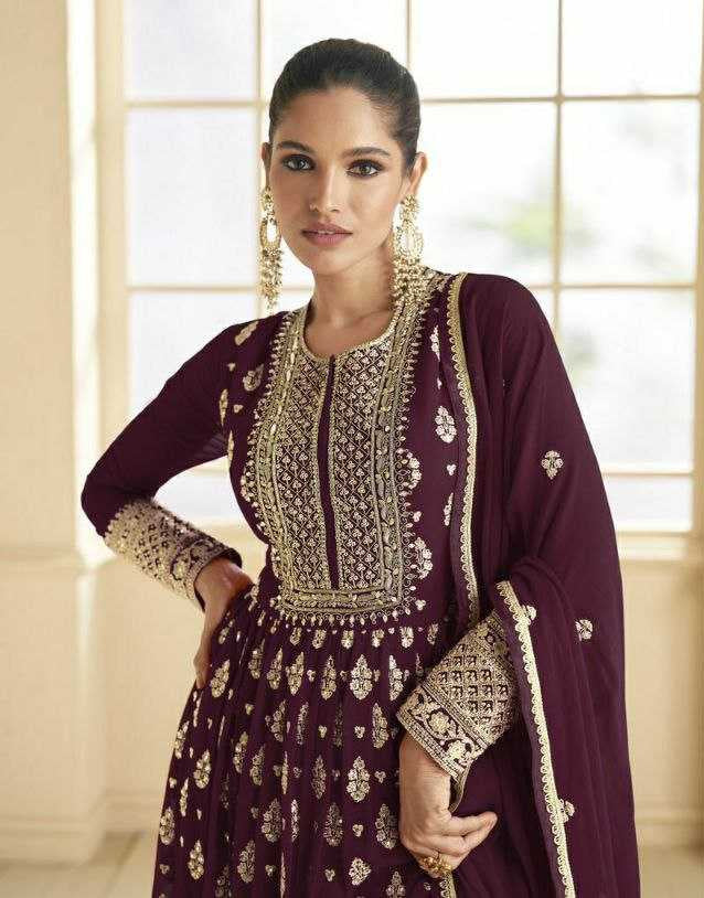 Woman in a maroon embroidered traditional outfit with gold details, standing in a bright room.