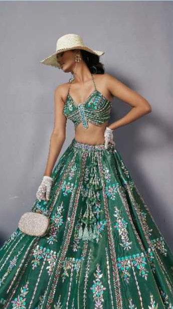 Woman in a green and silver lehenga with a white hat, posing against a light blue background.