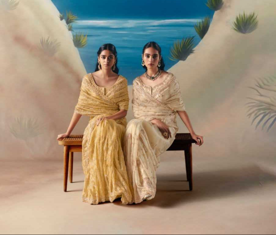 Two women in traditional attire sitting on a bench with a scenic background, featuring the brand 'Kimora Premium Chandani'.