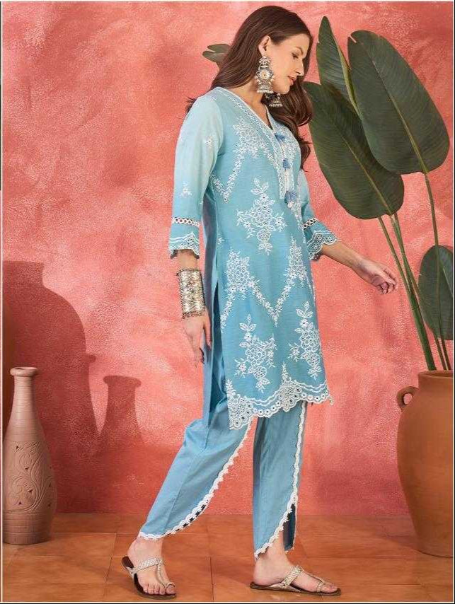 Woman wearing a light blue embroidered outfit against a pink wall with decorative vases and plants.