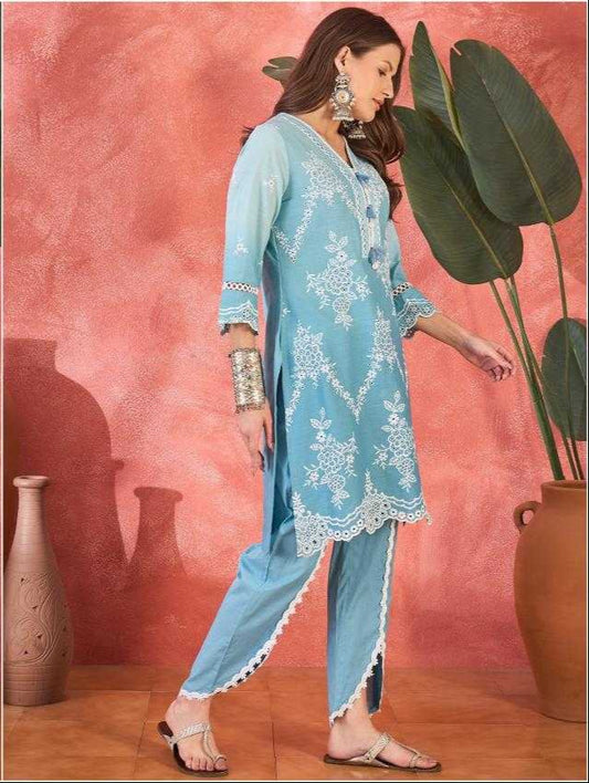 Woman wearing a light blue embroidered outfit against a pink wall with decorative vases and plants.