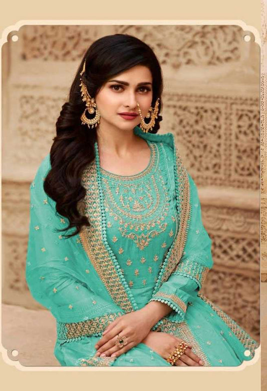 Woman in a traditional turquoise outfit with gold embroidery standing in front of an architectural background.
