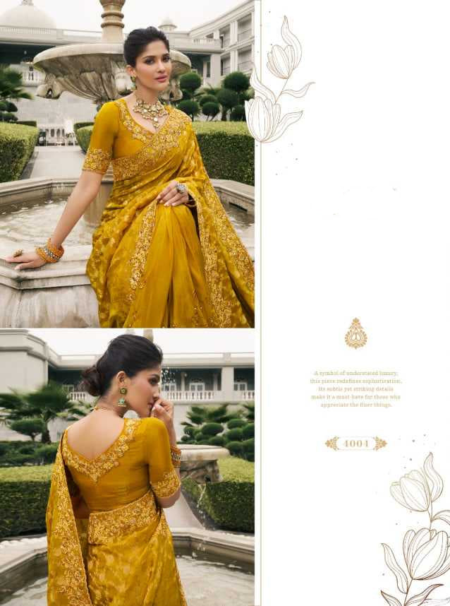 Woman in a yellow saree standing outdoors with a fountain and building in the background.