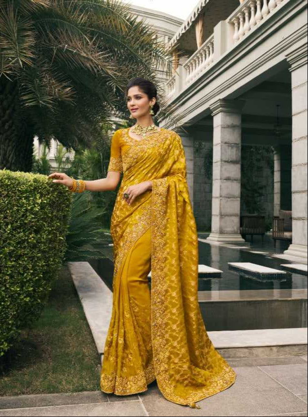 Woman in a yellow saree standing outdoors with a fountain and building in the background.