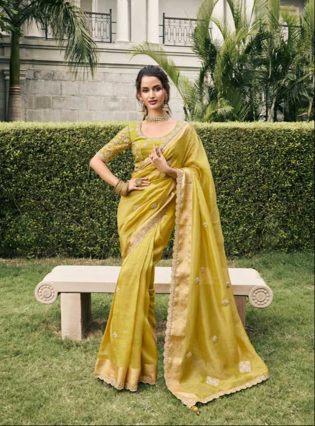 Woman in a yellow saree sitting on grass with a bench and hedge in the background