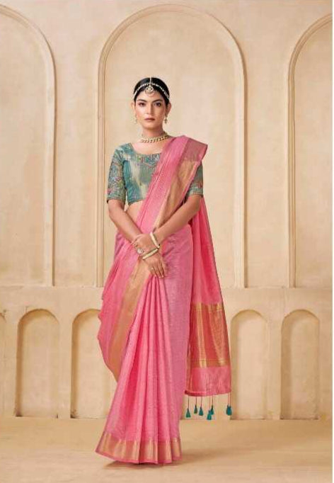 Woman in a green blouse and pink saree with gold border, standing against a beige architectural background.